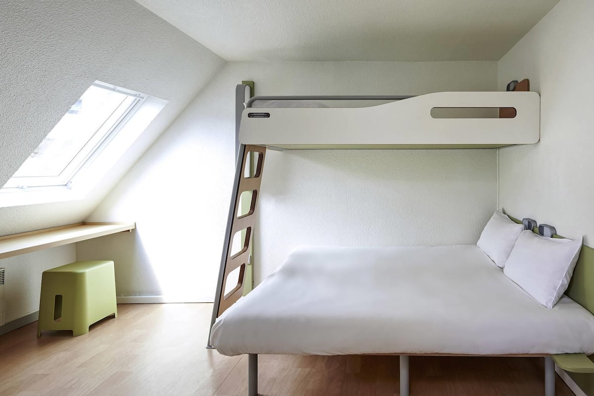Uma cama beliche com escrivaninha e um banquinho num quarto com teto inclinado e claraboia.