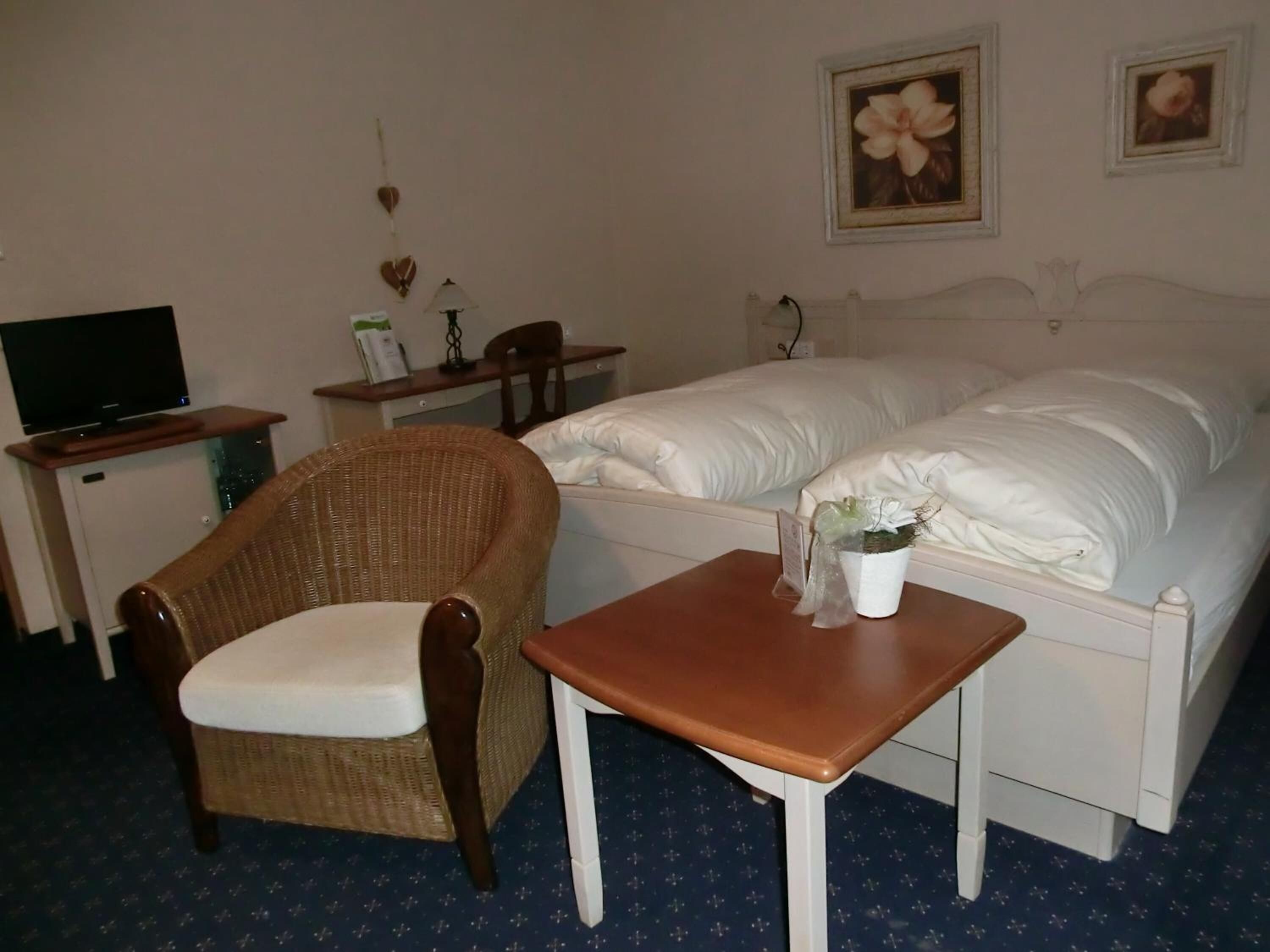Quarto de hotel com cama, uma cadeira, uma mesinha, televisão e duas fotos emolduradas na parede.