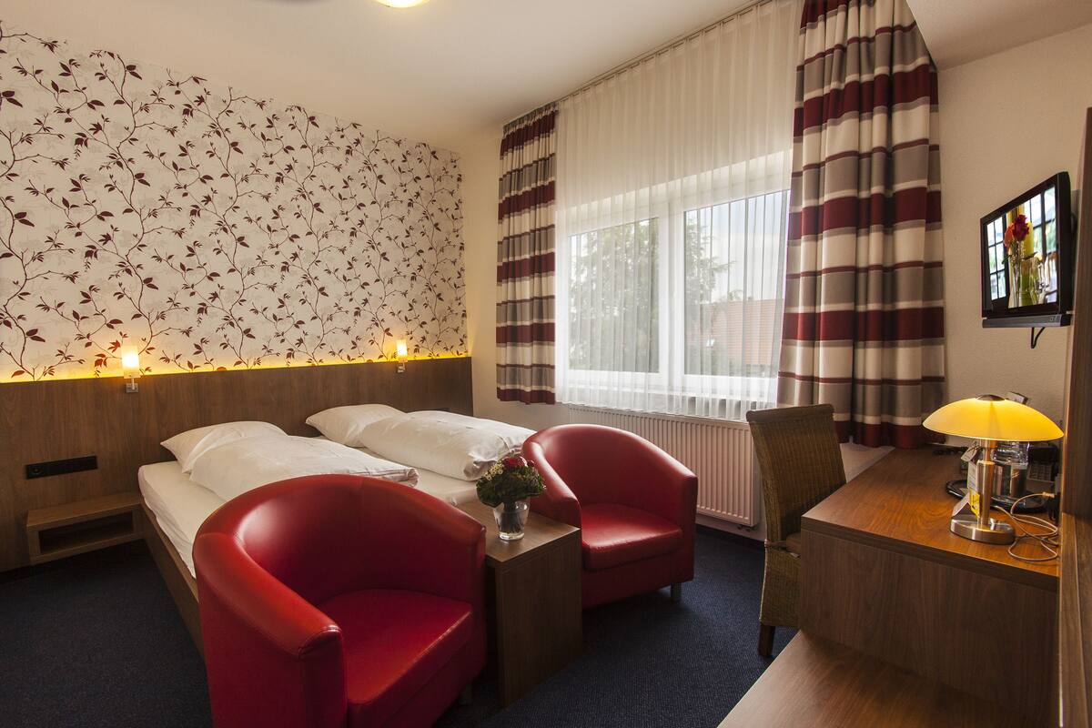 Habitación de hotel con dos camas, sillas rojas, un escritorio de madera, una lámpara y una pared con estampado floral.