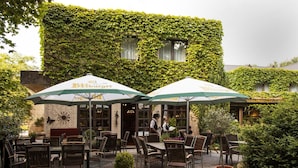 Un espace extérieur aménagé en terrasse, avec des meubles en osier et des parasols, un mur de briques recouvert de lierre, et une personne qui marche au loin.