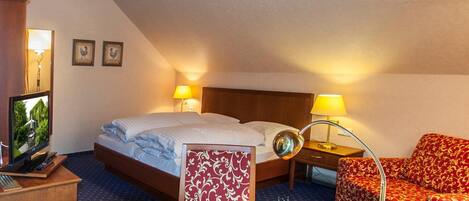Habitación de hotel con cama, dos mesitas de noche, un escritorio con computadora, una silla, un sofá y un jarrón decorativo con flores.