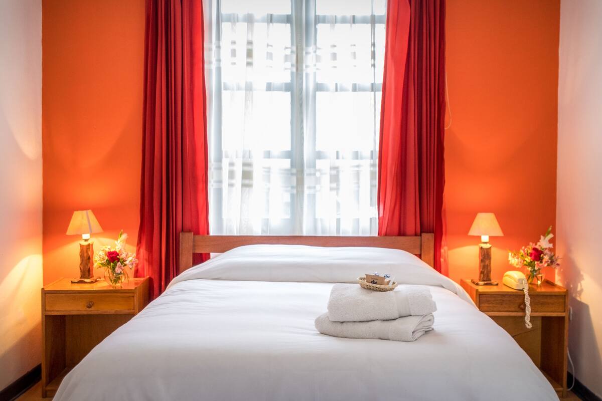 Ett hotellrum med en stor säng, röda gardiner, två sängbord med lampor och ett fönster med utsikt över byggnader.