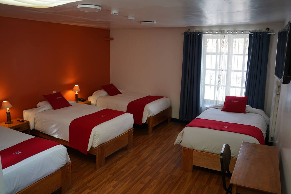 Ett hotellrum med tre sängar, trägolv, ett skrivbord och ett fönster med gardiner.