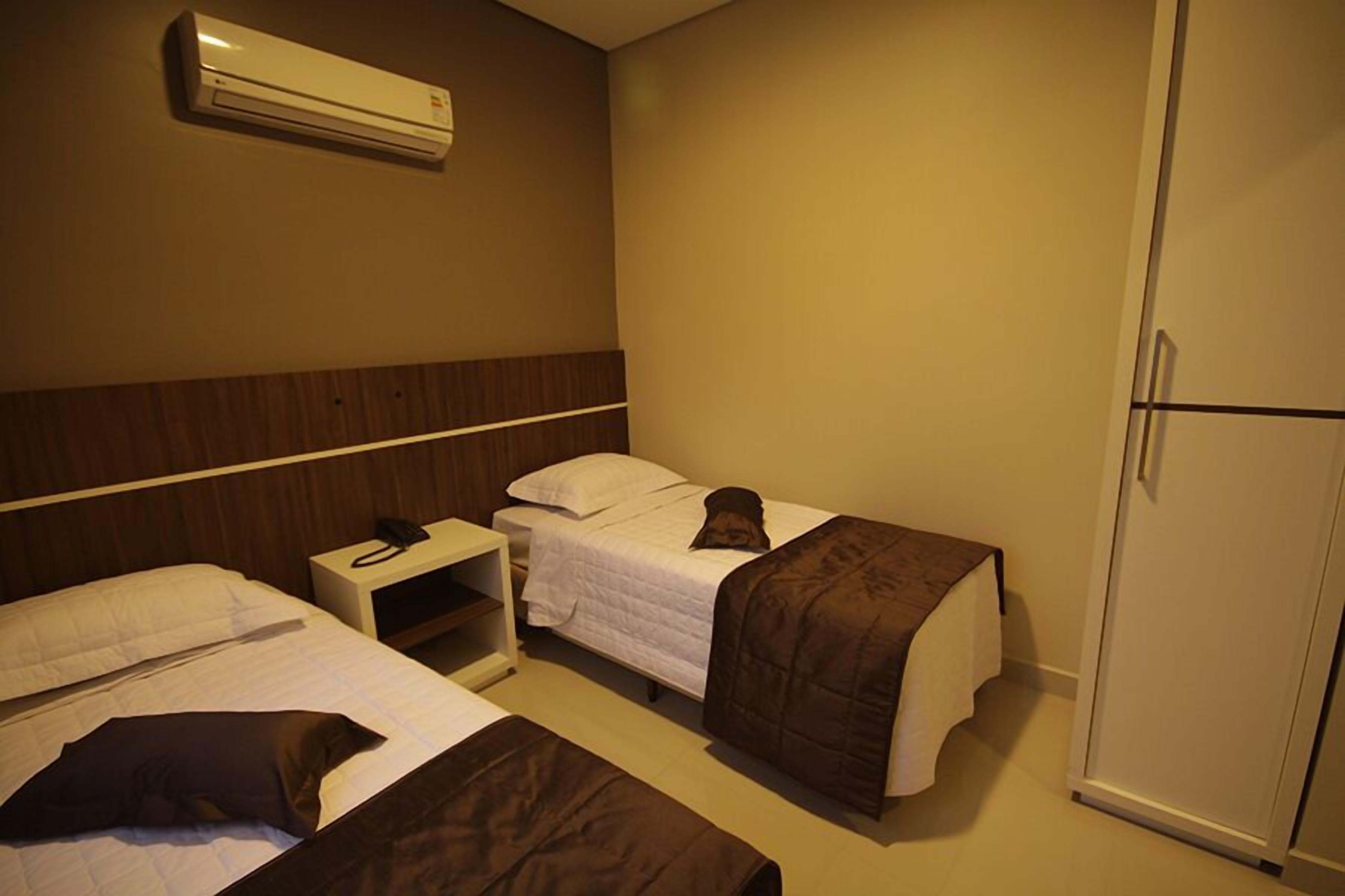 A hotel room with two beds, a wardrobe, a bedside table with a phone, and an air conditioning unit.