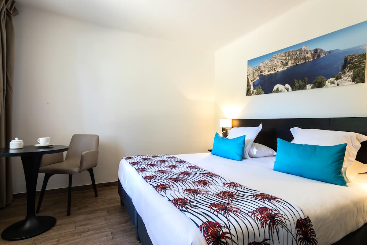 Una habitación de hotel con una cama, un escritorio con una silla, una mesa con una taza y una tetera, y un cuadro en la pared.