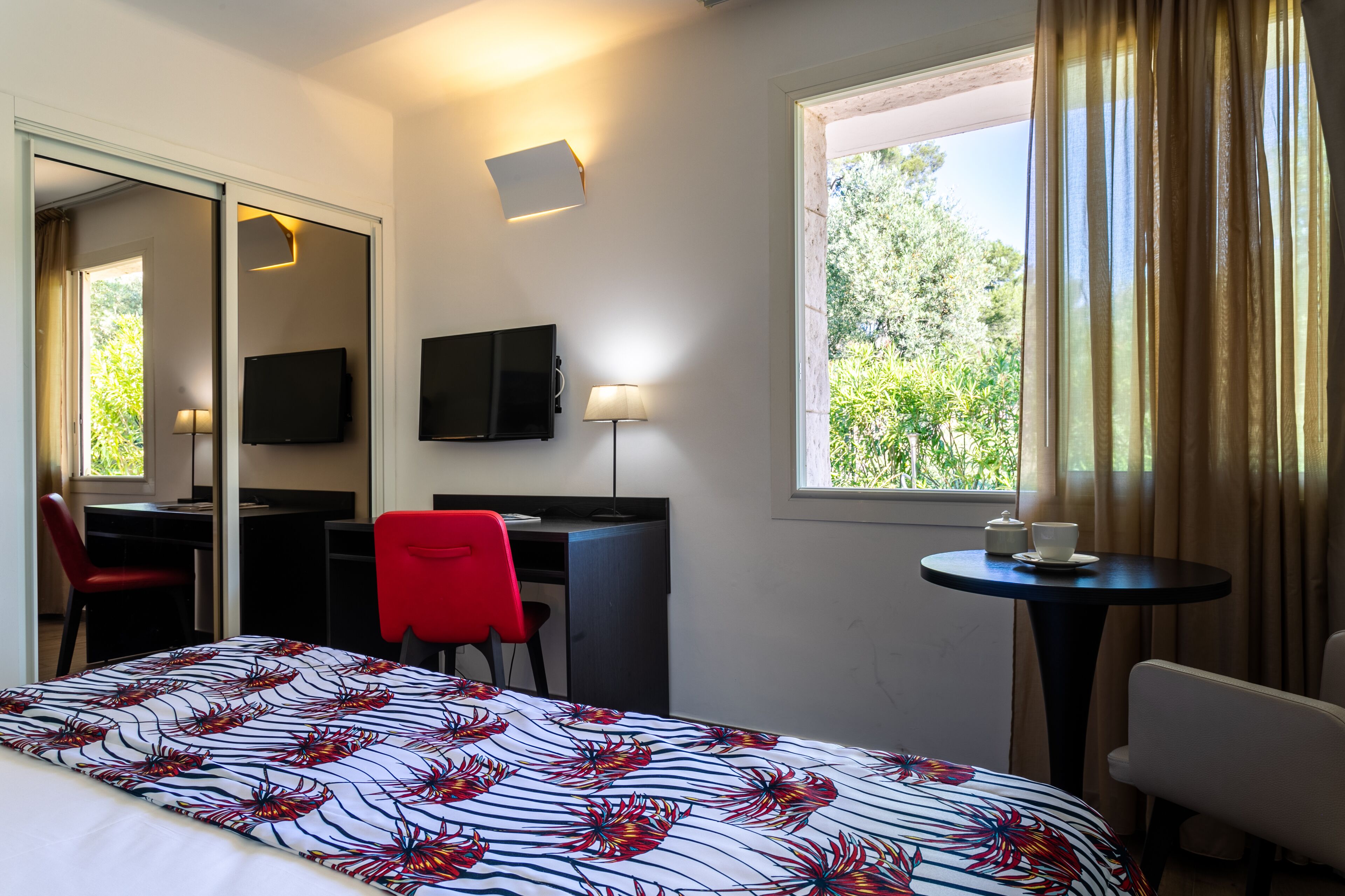 Hotelkamer met een bed, een bureau, een stoel, een televisie, een lamp, een klein tafeltje met een kopje en schotel, en een raam met gordijnen.