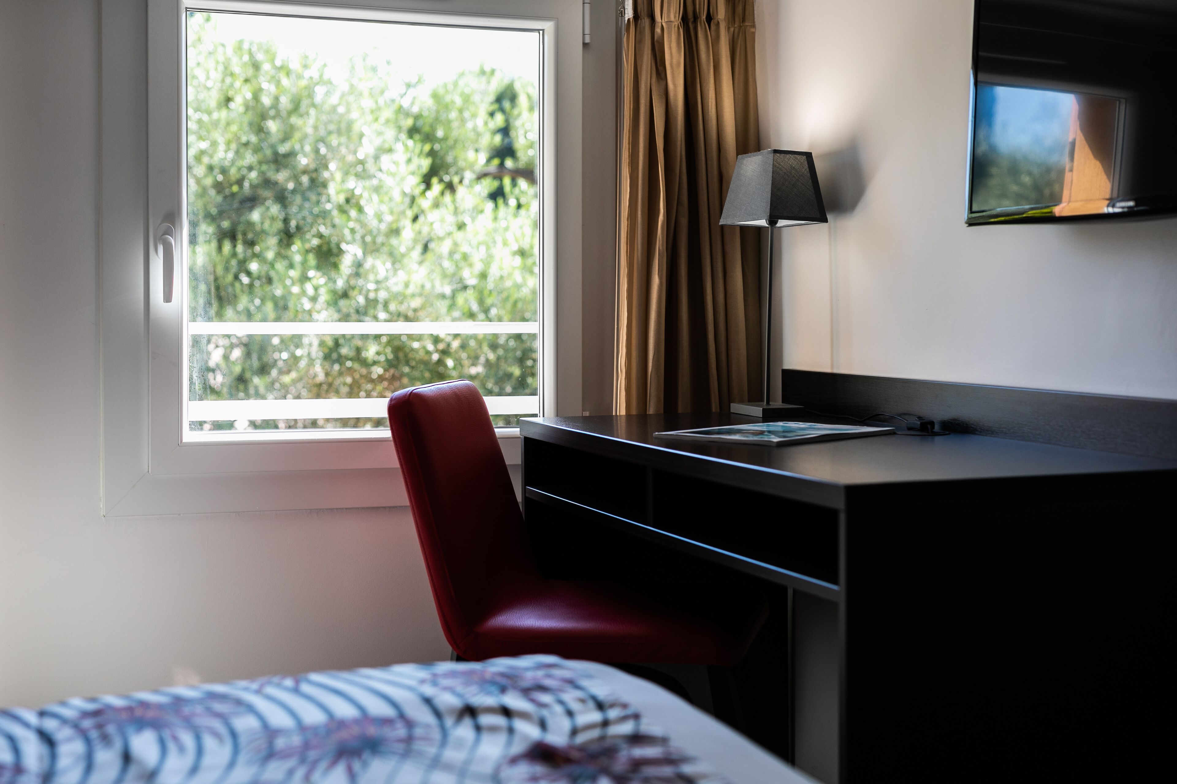 A hotel room with a bed, a desk, a chair, a TV, and a window with a view of trees.