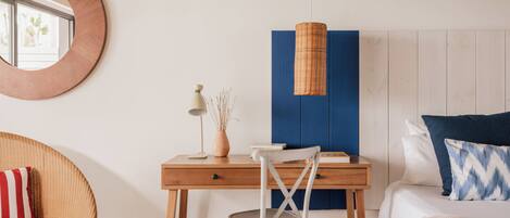 A room with a wooden desk, a chair, a bed with pillows, a round mirror, and a hanging light fixture.