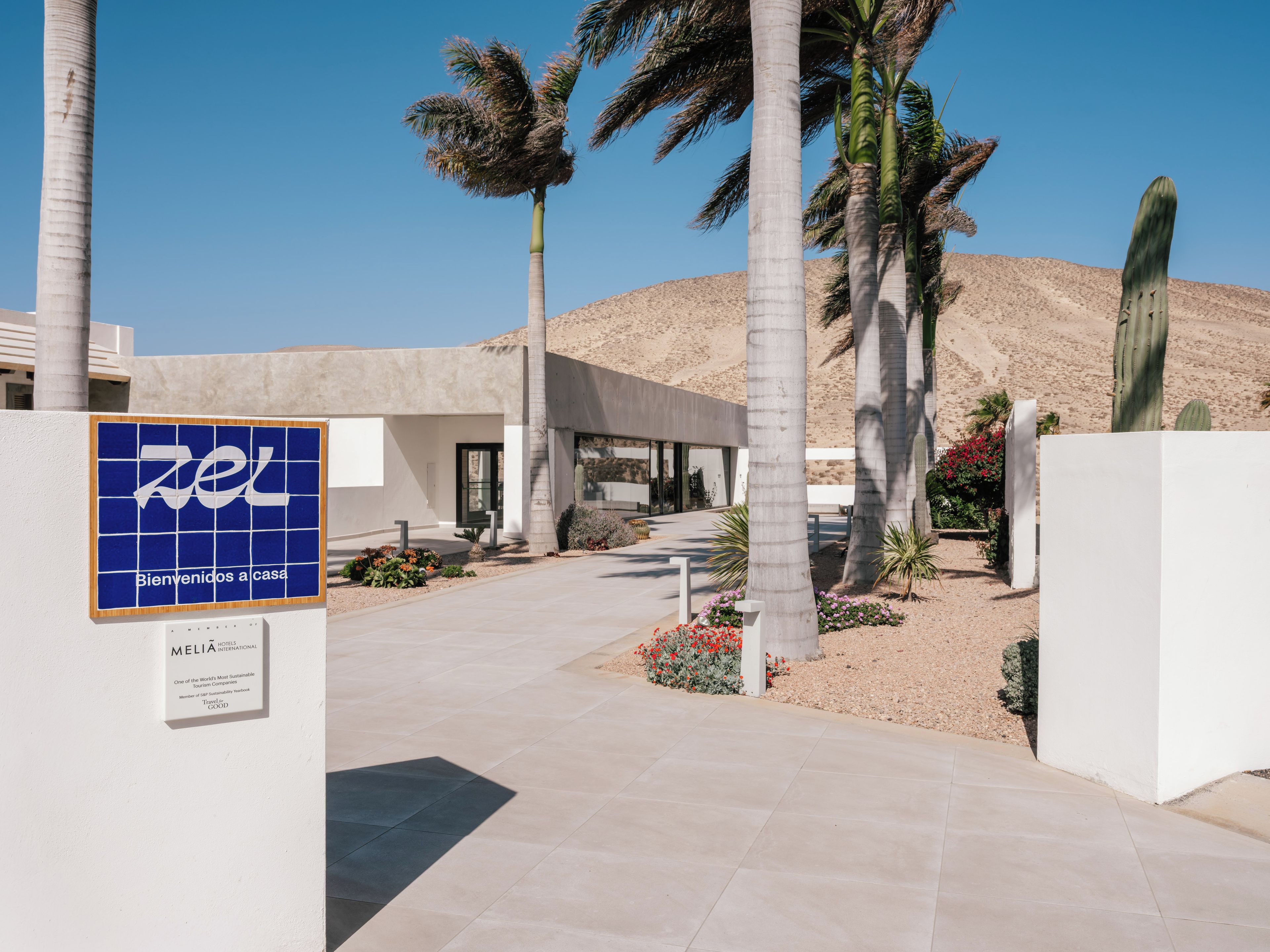 A hotel entrance with a blue sign, palm trees, and a desert landscape.