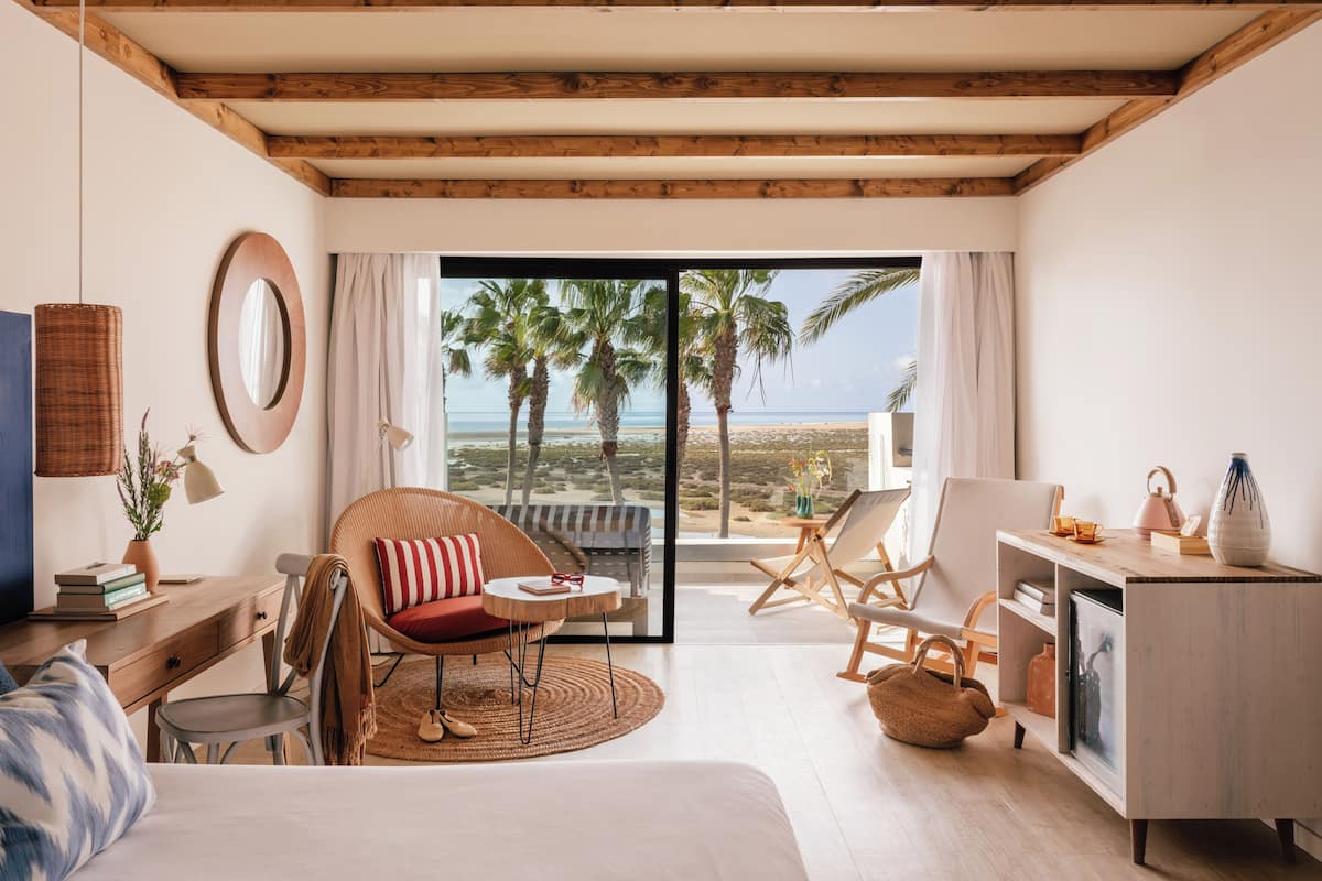A room with a bed, a desk, a chair, a small table, a sofa, a large window with a view of palm trees and the beach, and a wooden ceiling.