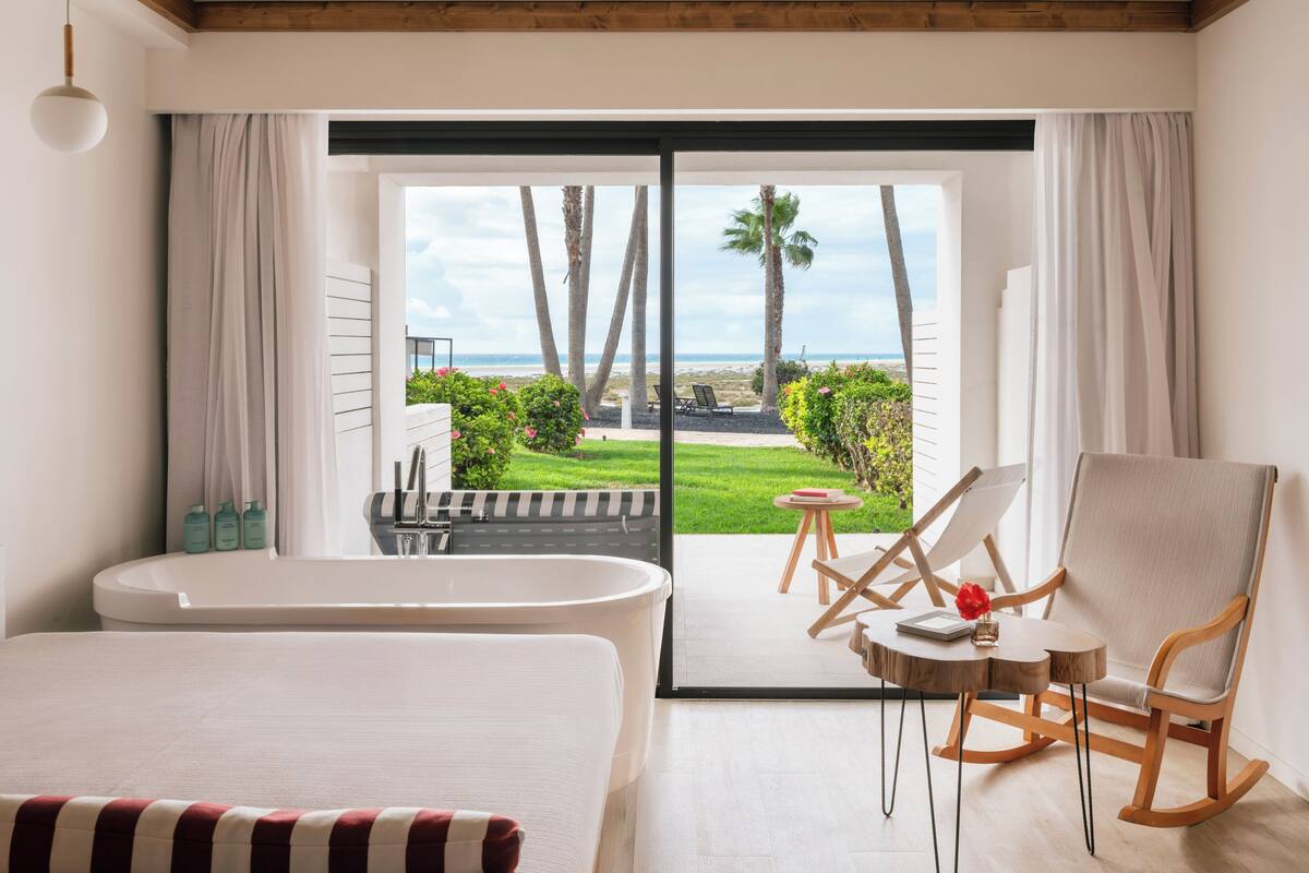 A hotel room with a large bed, a bathtub, a rocking chair, and a view of the beach and palm trees.