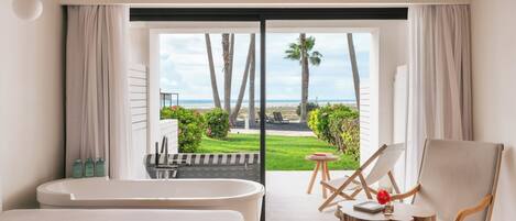 A hotel room with a large bed, a bathtub, a rocking chair, and a view of the beach and palm trees.