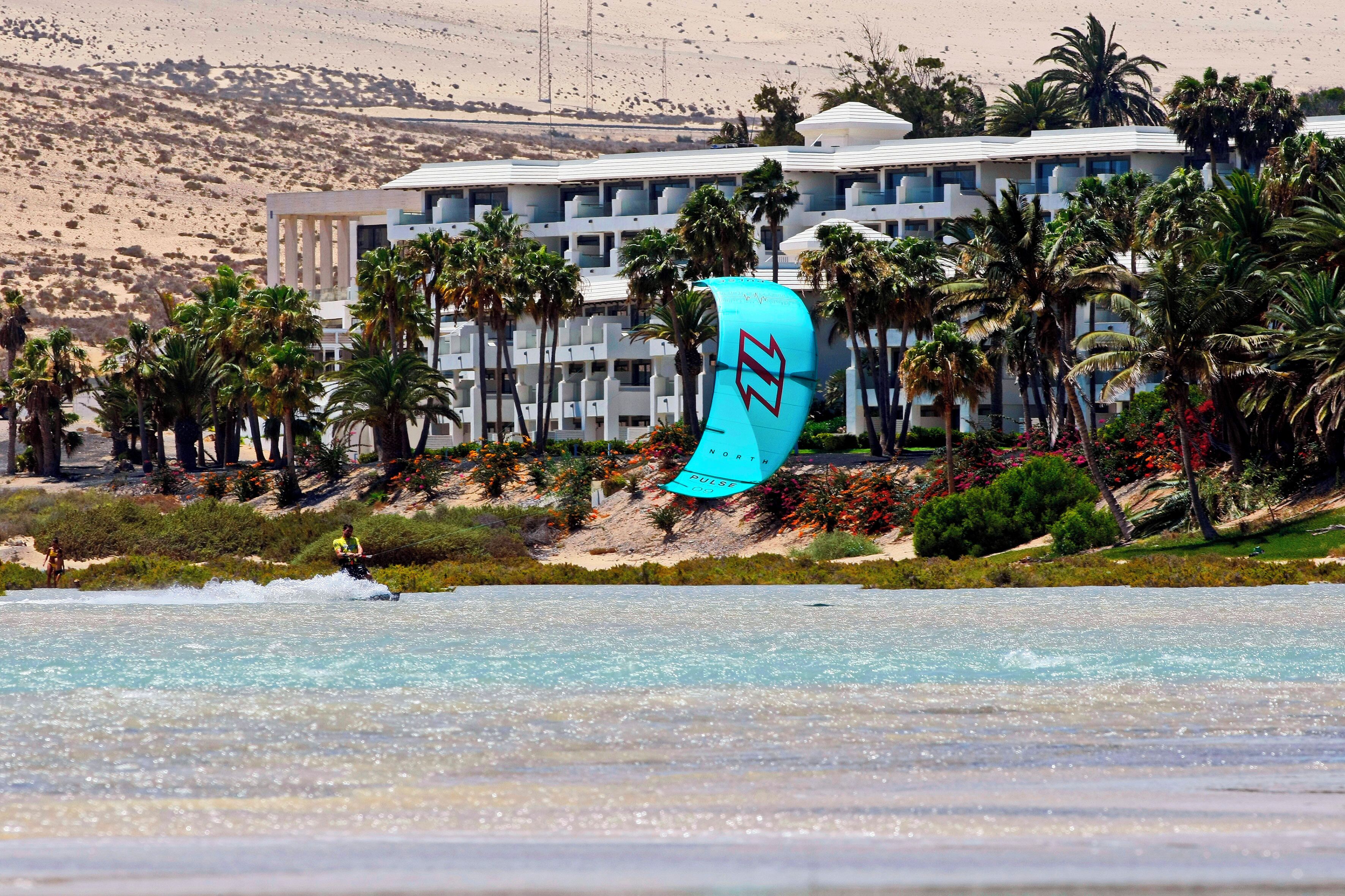 Un hotel frente a la playa con piscina, palmeras y una persona practicando kitesurf.