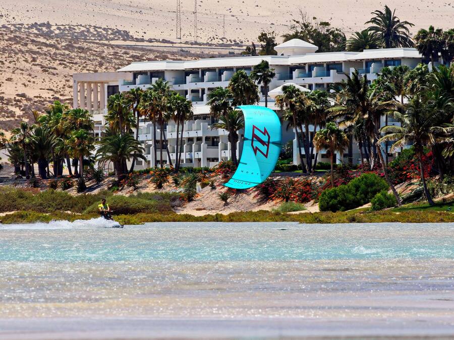 A beachfront hotel with a pool, palm trees, and a person kiteboarding.
