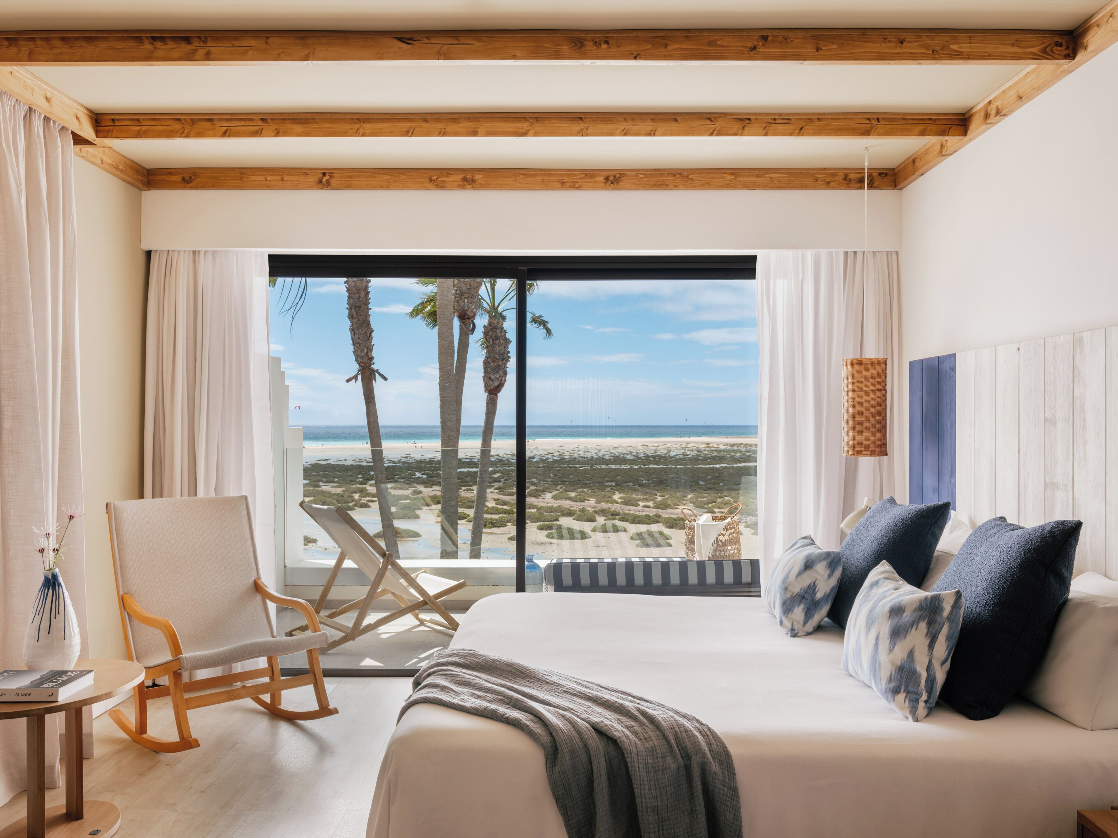 Une chambre avec un grand lit, un fauteuil à bascule, une vue sur la plage et un plafond en bois.