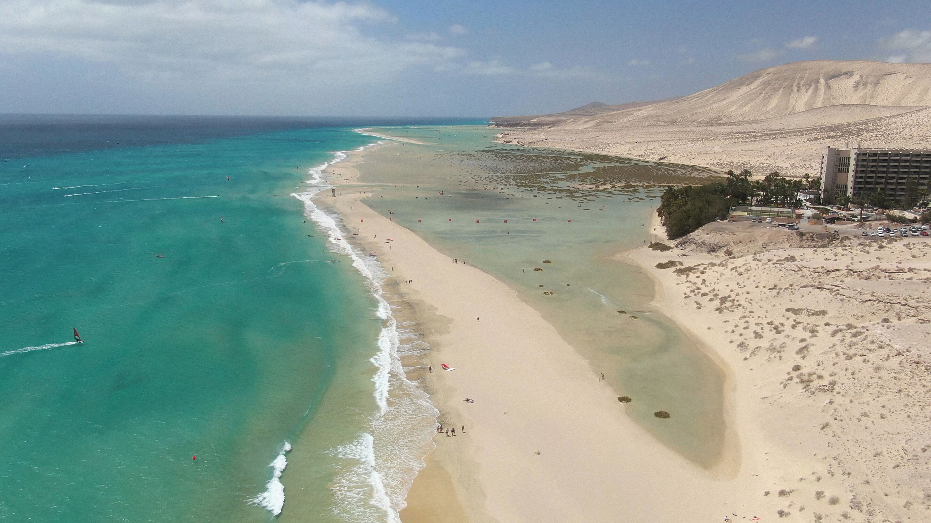 A coastal beach with clear turquoise waters, a sandy shore, and a desert landscape in the background.