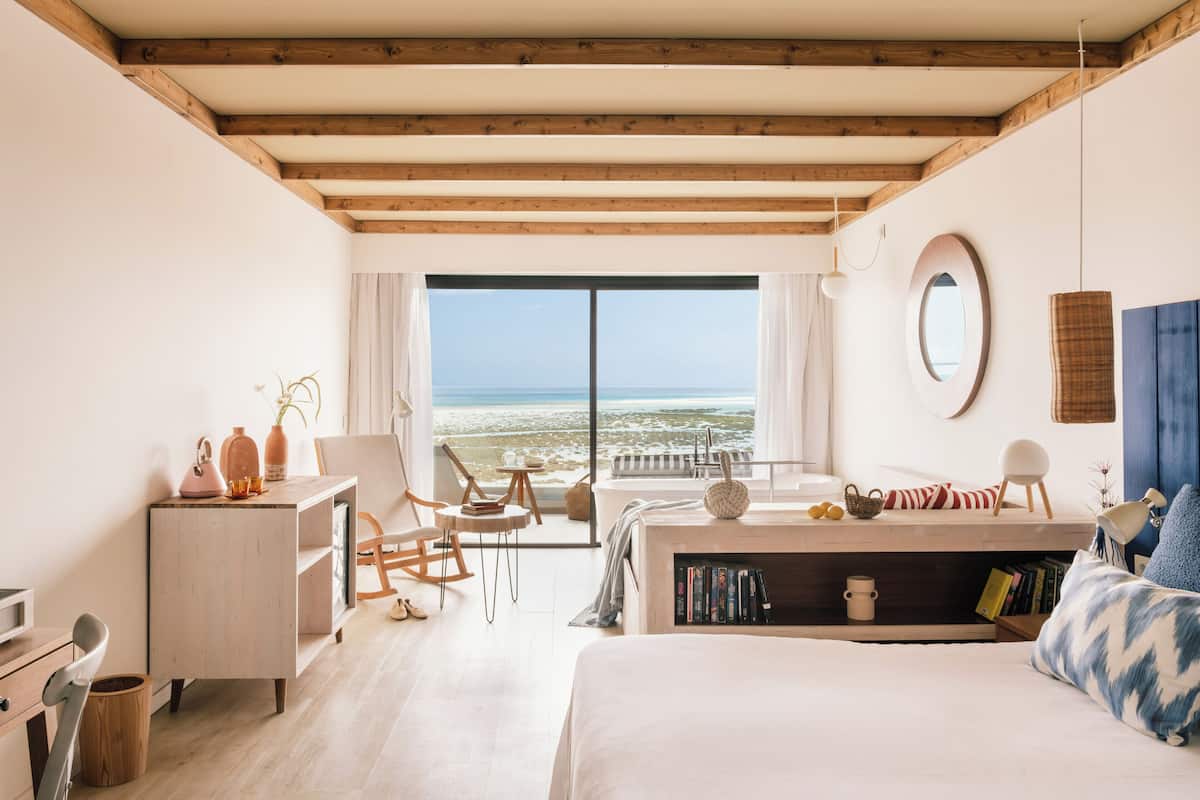 A bedroom with a large bed, a desk, a chair, a bookshelf, and a view of the beach.