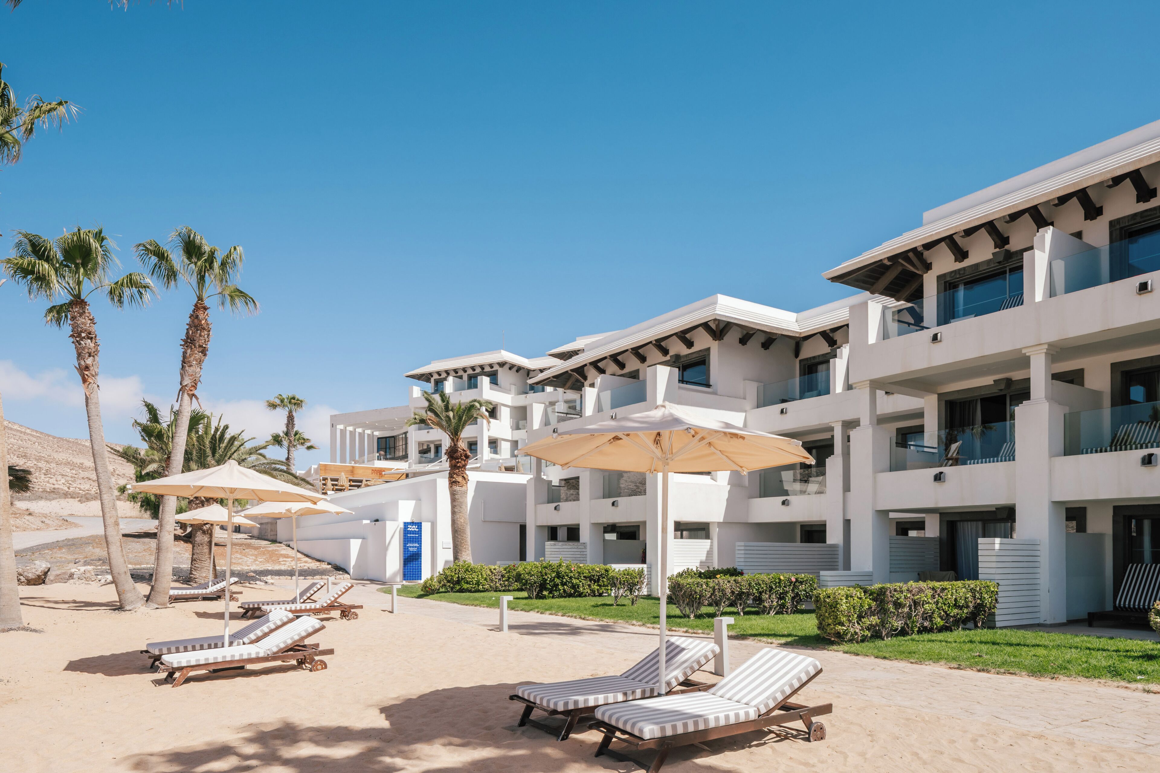 A modern hotel with multiple floors, outdoor seating with umbrellas, and palm trees.