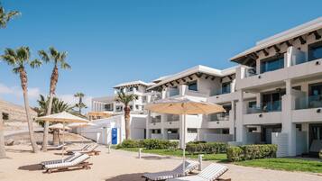 Un hotel moderno con varios pisos, mesas exteriores con sombrillas y palmeras.