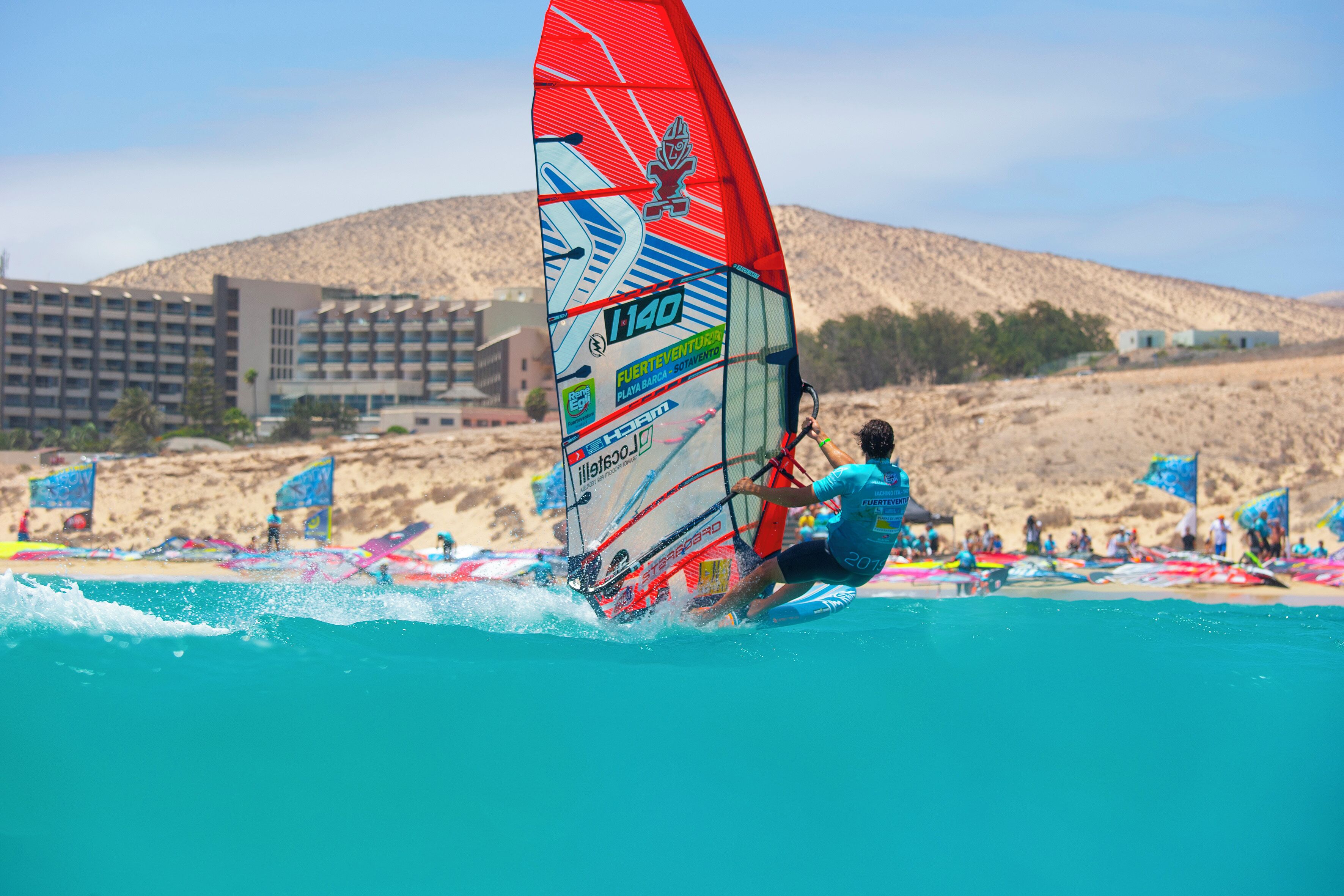 Una persona practica windsurf en una playa, con un edificio al fondo.