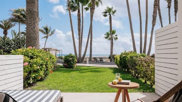Un patio con un cojín a rayas, una silla de madera, una pequeña mesa redonda con un libro y fruta, y vista al océano con palmeras.