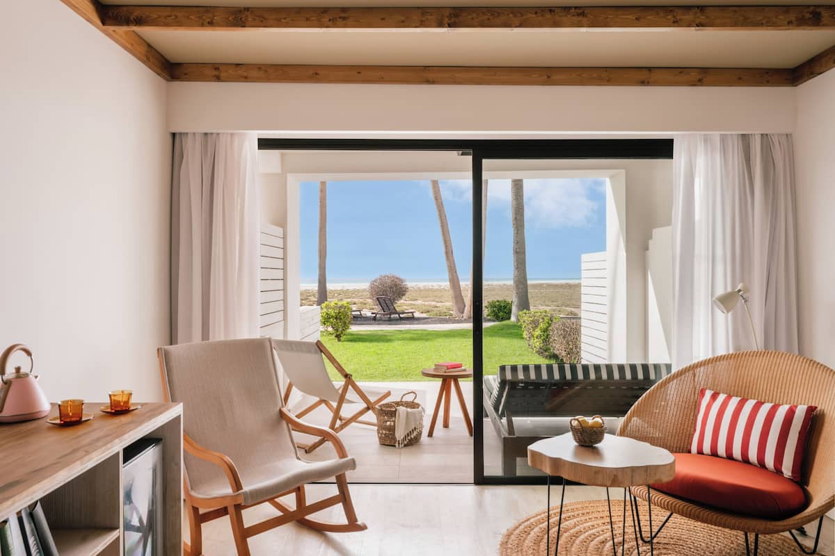 A room with a wooden ceiling, a sliding glass door to an outdoor area, a rocking chair, a sofa, a small table, and a bookshelf.
