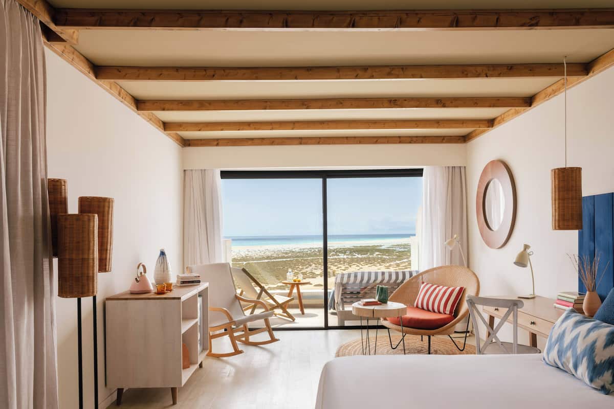 A room with a bed, a desk, a chair, a small table, and a view of the beach.