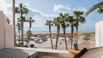 Un balcón con una cama, una silla y vistas a la playa y palmeras.