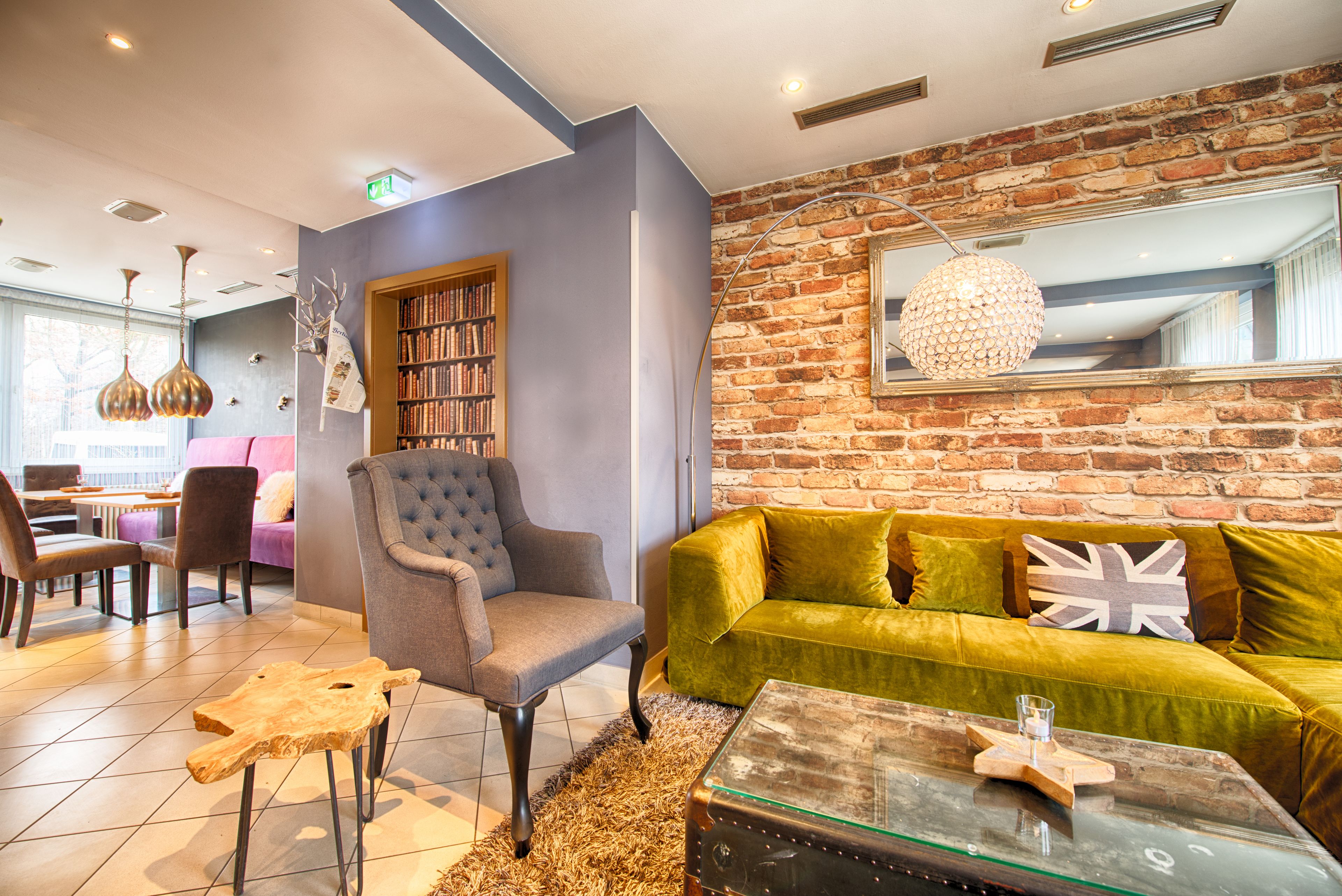 Un espacio lounge moderno con pared de ladrillo, un sofá verde, un sillón capitoné, una mesa de centro de vidrio y una estantería.