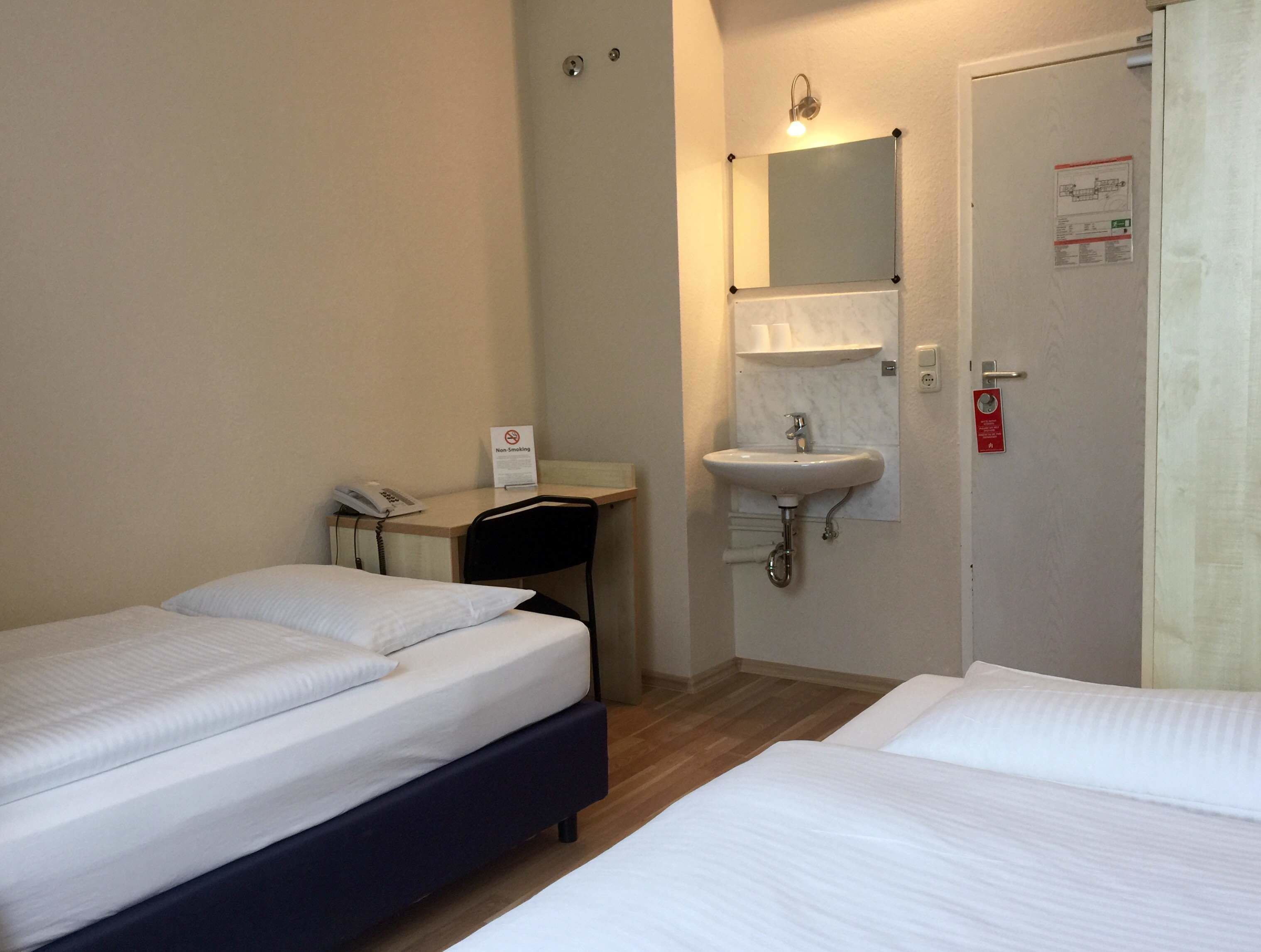A hotel room with two beds, a desk with a telephone, a non-smoking sign, a sink, and a door with a fire safety sign.