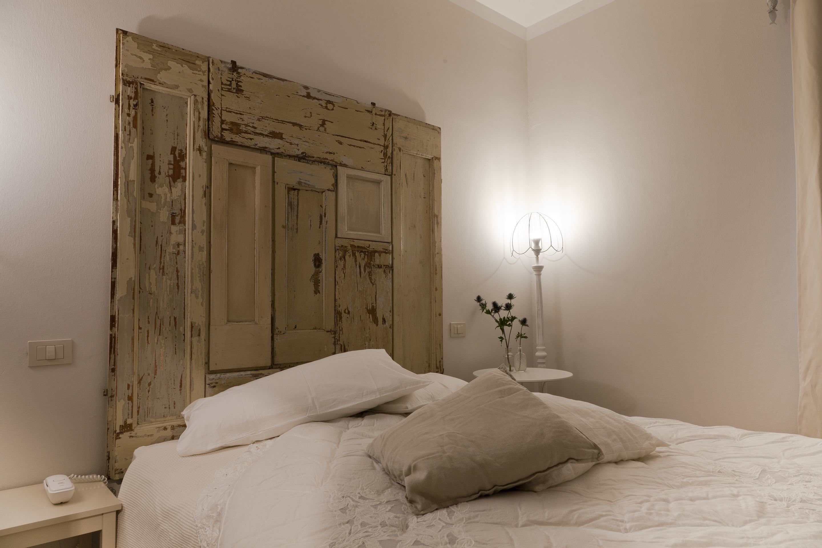 Une chambre à coucher avec un lit, une table de chevet, une lampe et un mur orné d’une porte ancienne.