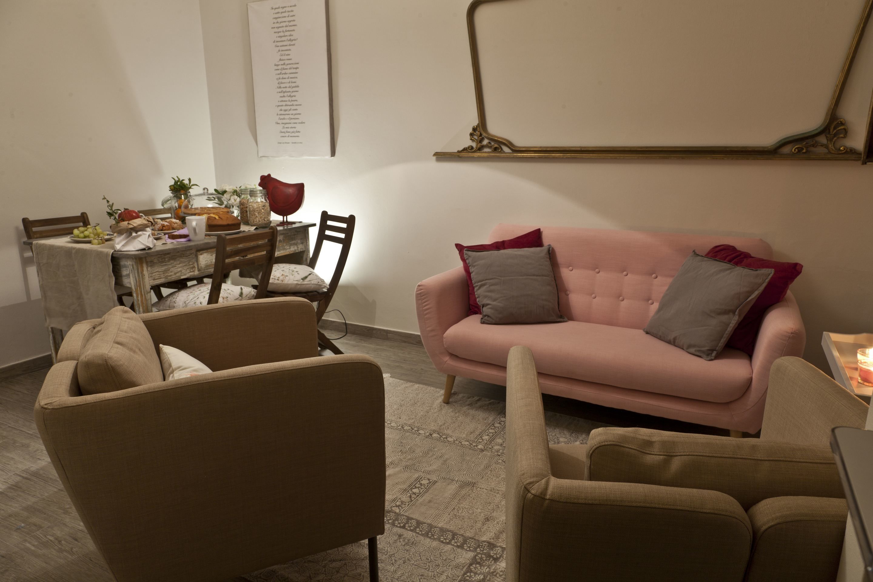 Un salon avec un canapé rose, des fauteuils beige, une table à manger dressée avec de la nourriture et des boissons, et un miroir décoratif accroché au mur.