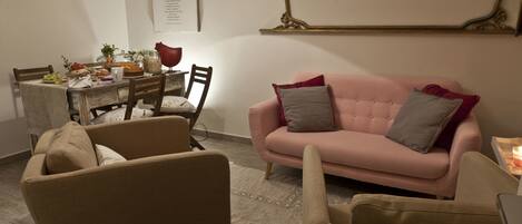 A living room with a pink sofa, beige armchairs, a dining table with food and drinks, and a decorative mirror on the wall.