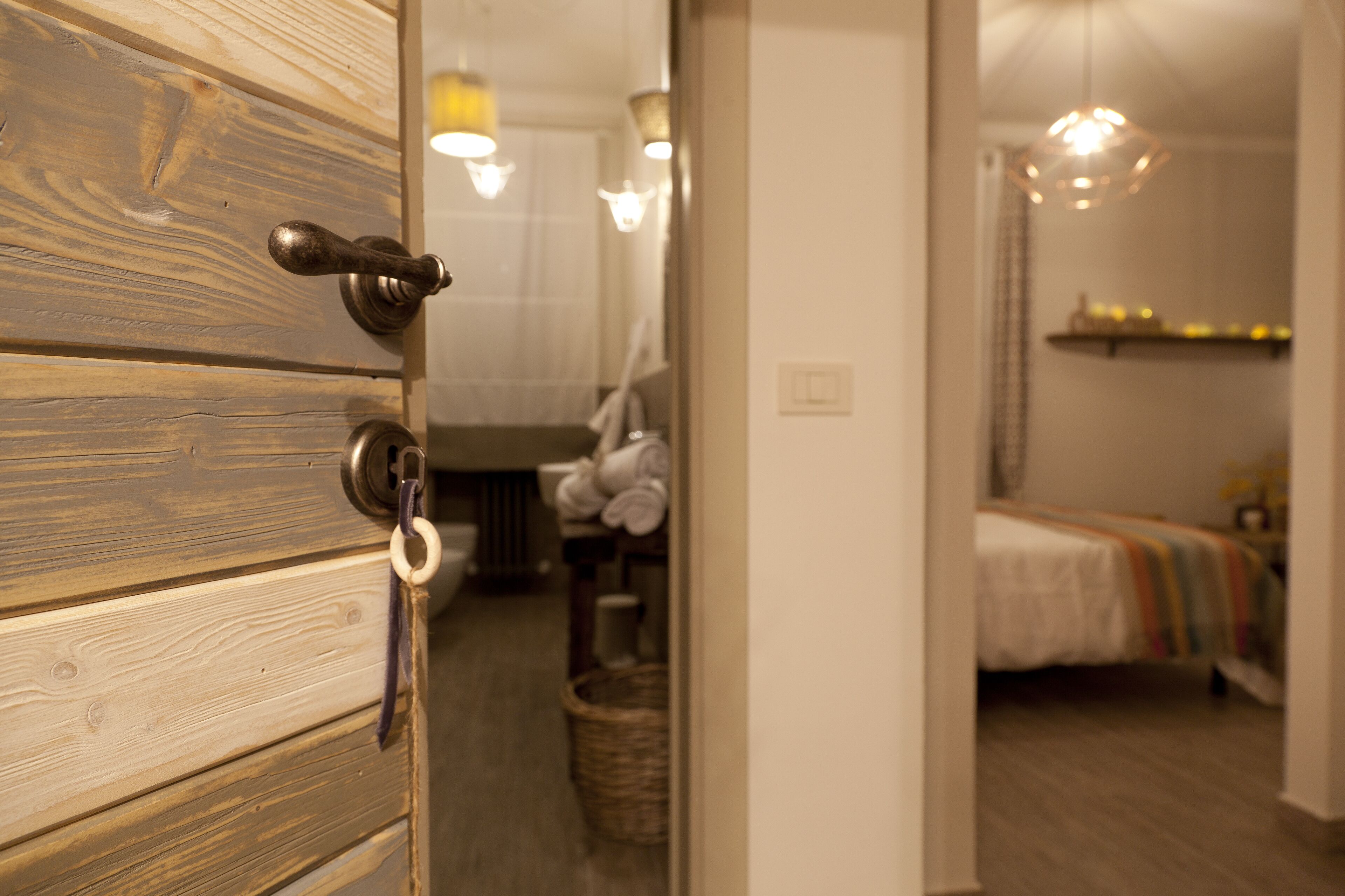 A wooden door with a keyhole and a ring, leading into a room with a bed, a hanging light fixture, and a basket on the floor.