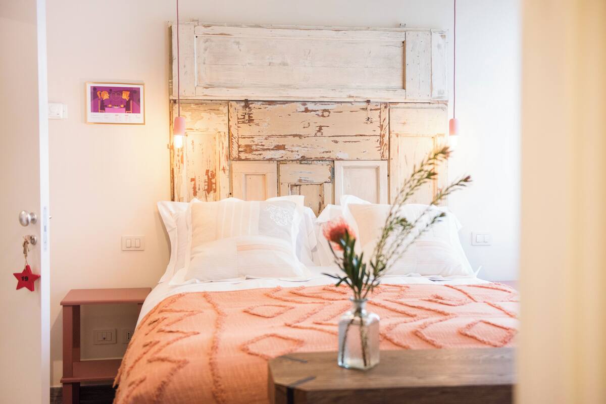 A bedroom with a rustic wooden headboard, a pink patterned bedspread, and a vase with flowers on a nightstand.