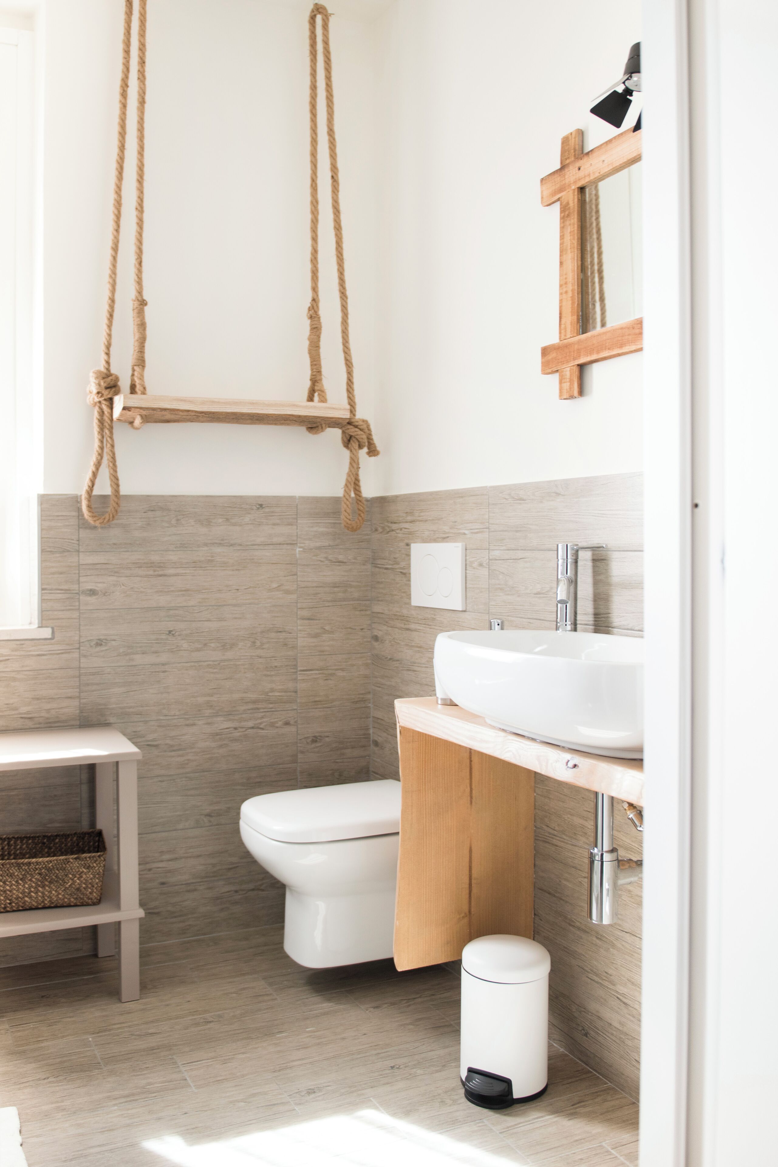 Un baño con tocador de madera, inodoro blanco y estante colgante de cuerda.