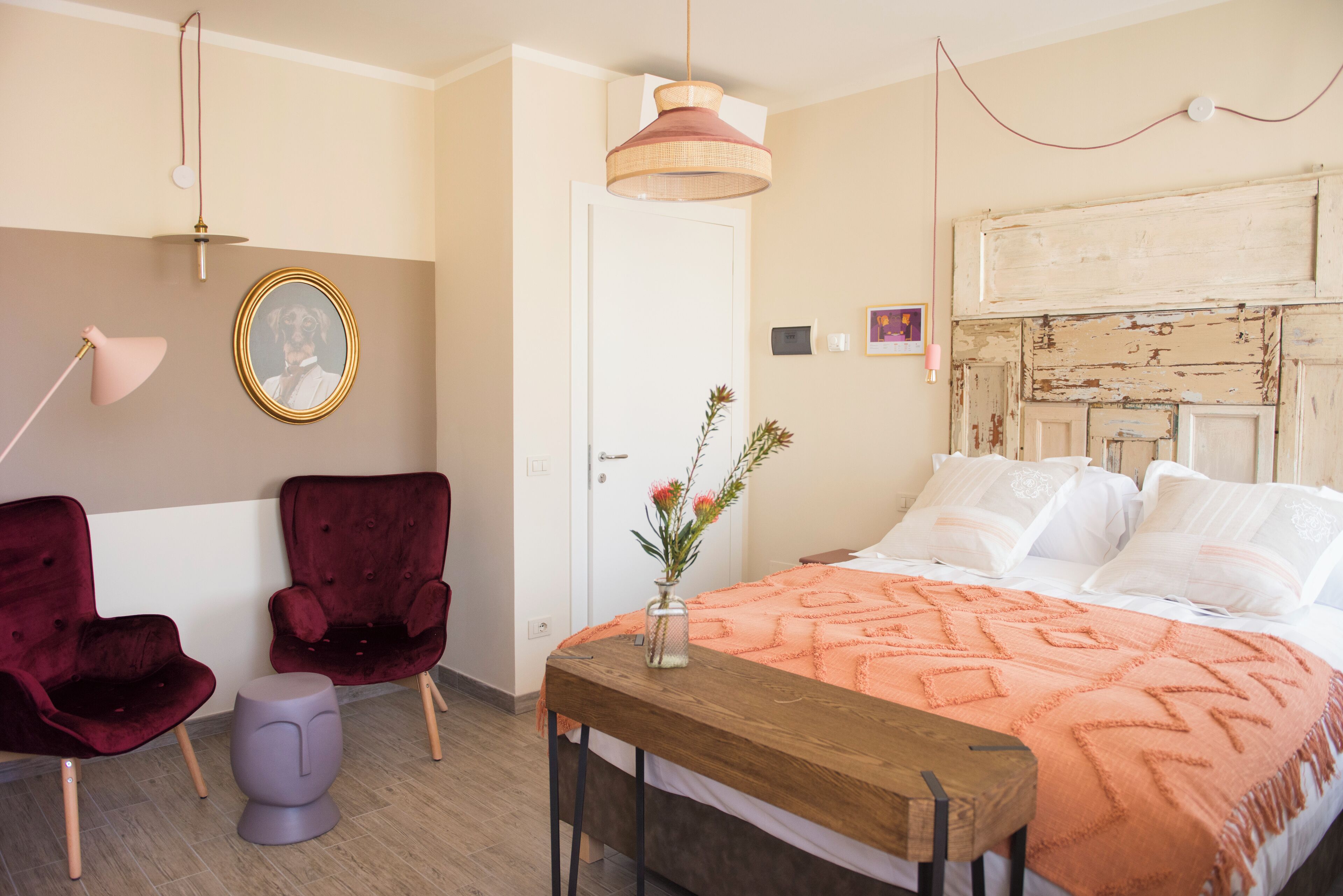 Een slaapkamer met een bed, twee stoelen, een tafel, een lamp, een spiegel en een muur met een afbeelding.