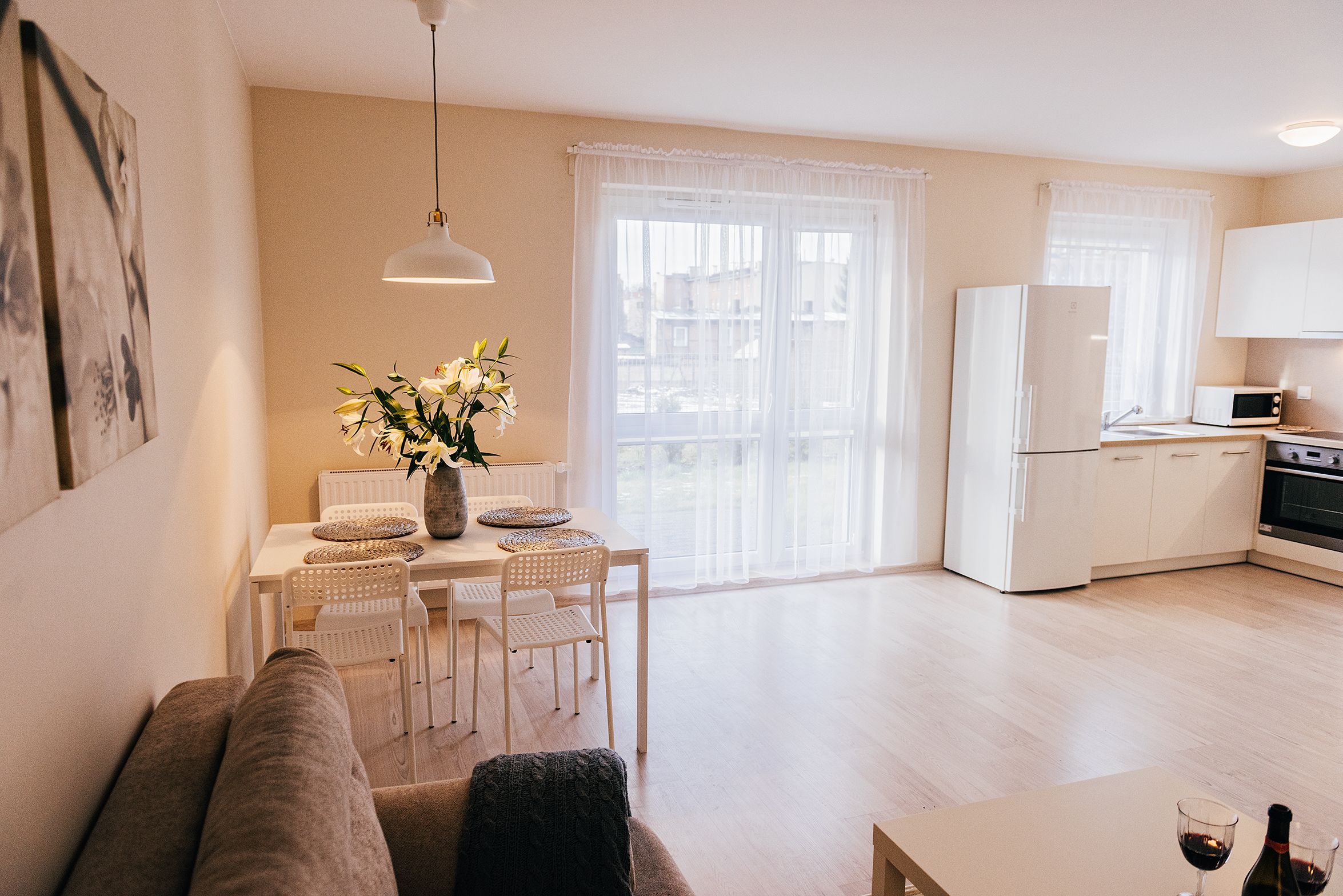 A modern living room with a sofa, dining table, and chairs. There is a kitchen area with a refrigerator, microwave, and oven. A large window with sheer curtains allows natural light into the room.