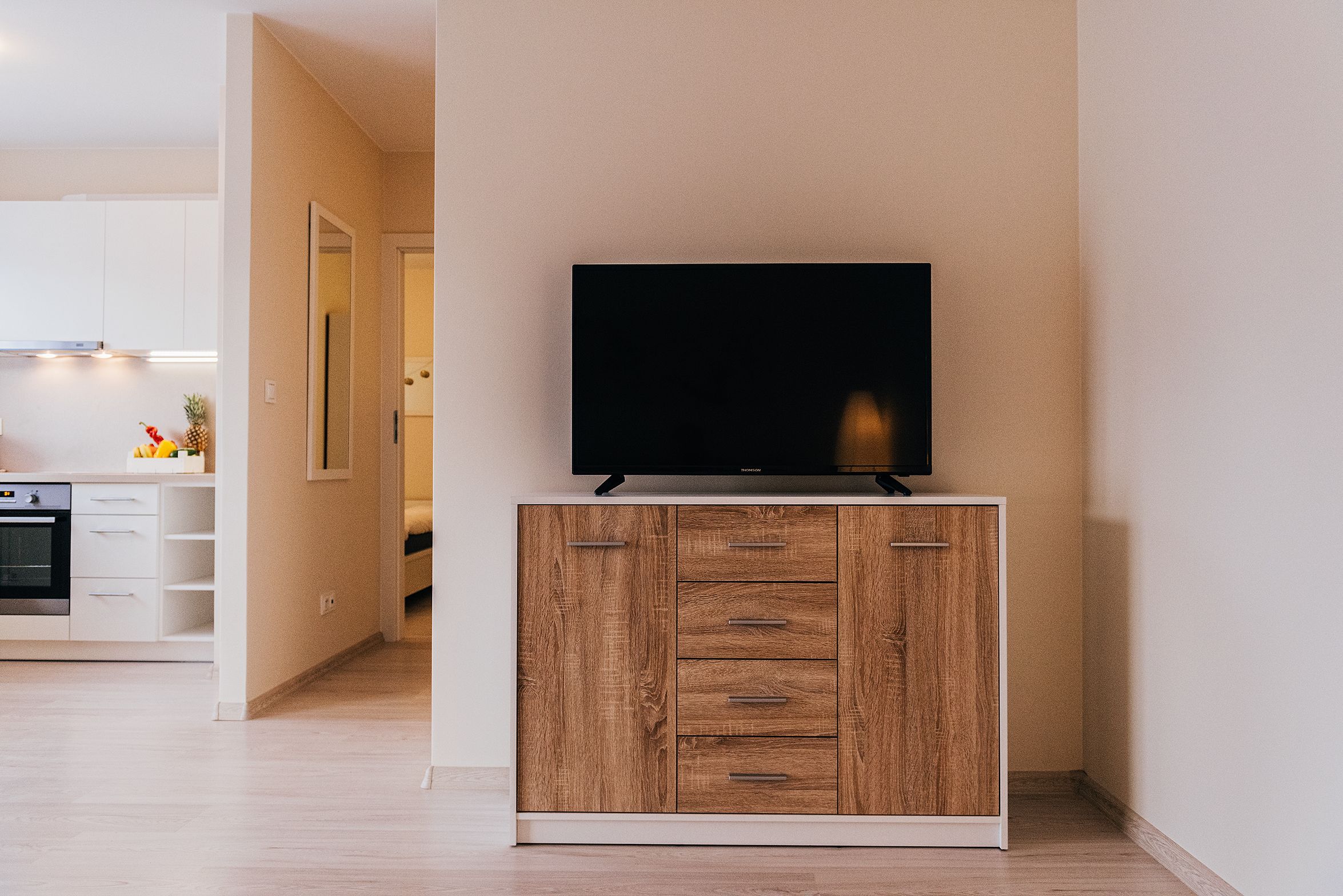 Een moderne keuken met witte kasten, een houten kast met lades, en een flatscreen televisie erboven.