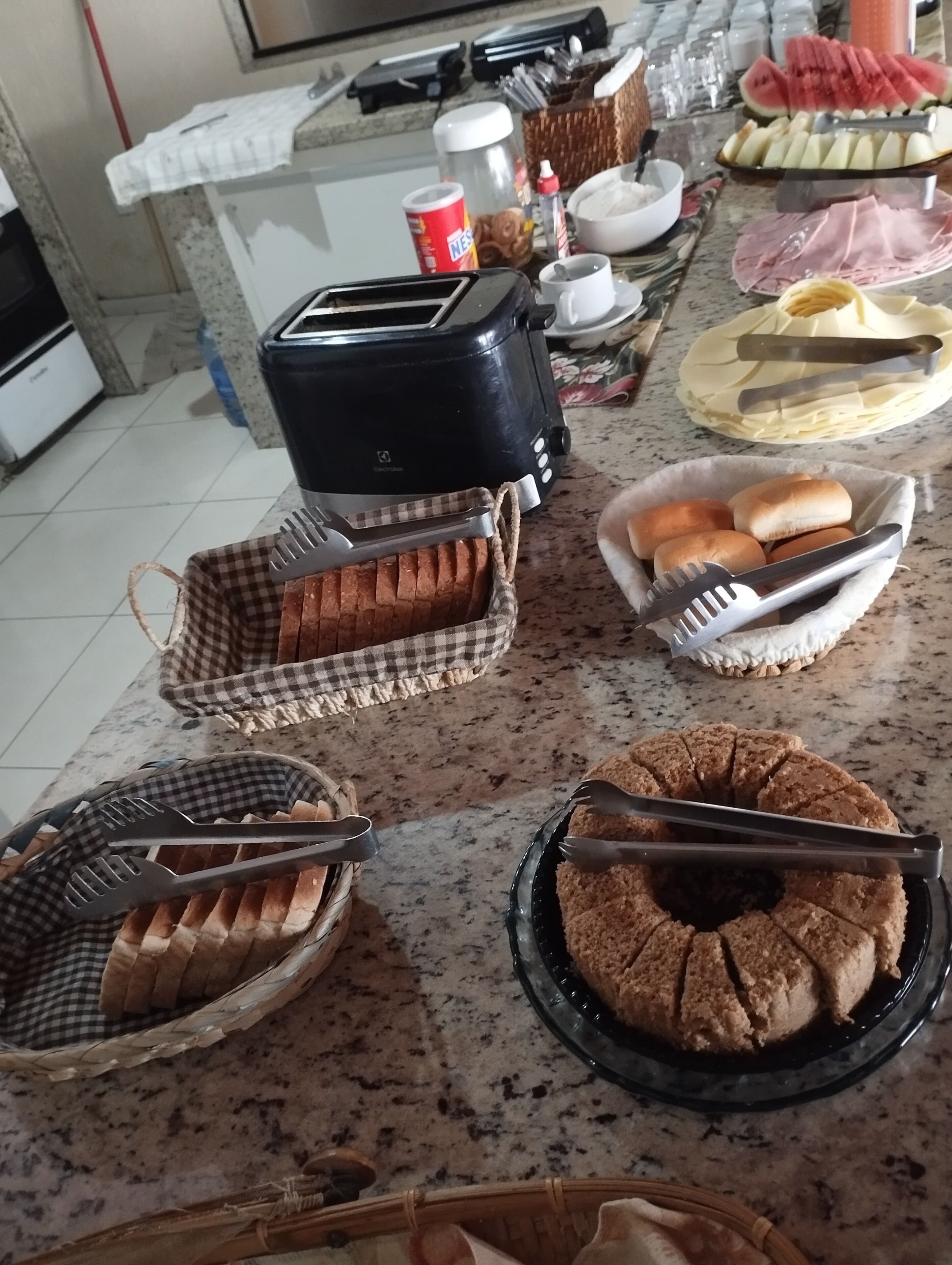 A kitchen counter with various food items and utensils, including a toaster, a bowl of bread, and a plate of sliced fruit.