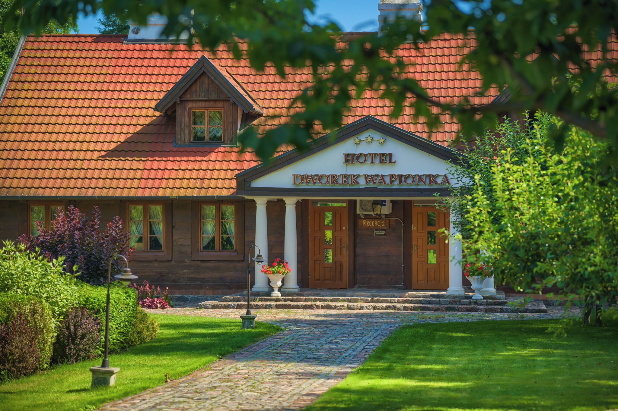 A hotel with a red-tiled roof, a wooden facade, and a welcoming entrance with a sign reading "HOTEL DWOREK WAPIONKA."