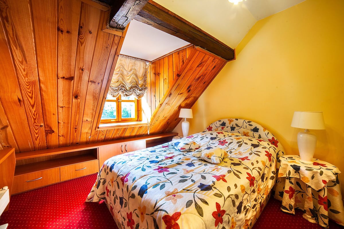 Une chambre avec un plafond en pente, des murs en bois, un lit avec une literie à motifs floraux, deux tables de chevet avec des lampes et une fenêtre avec des rideaux.