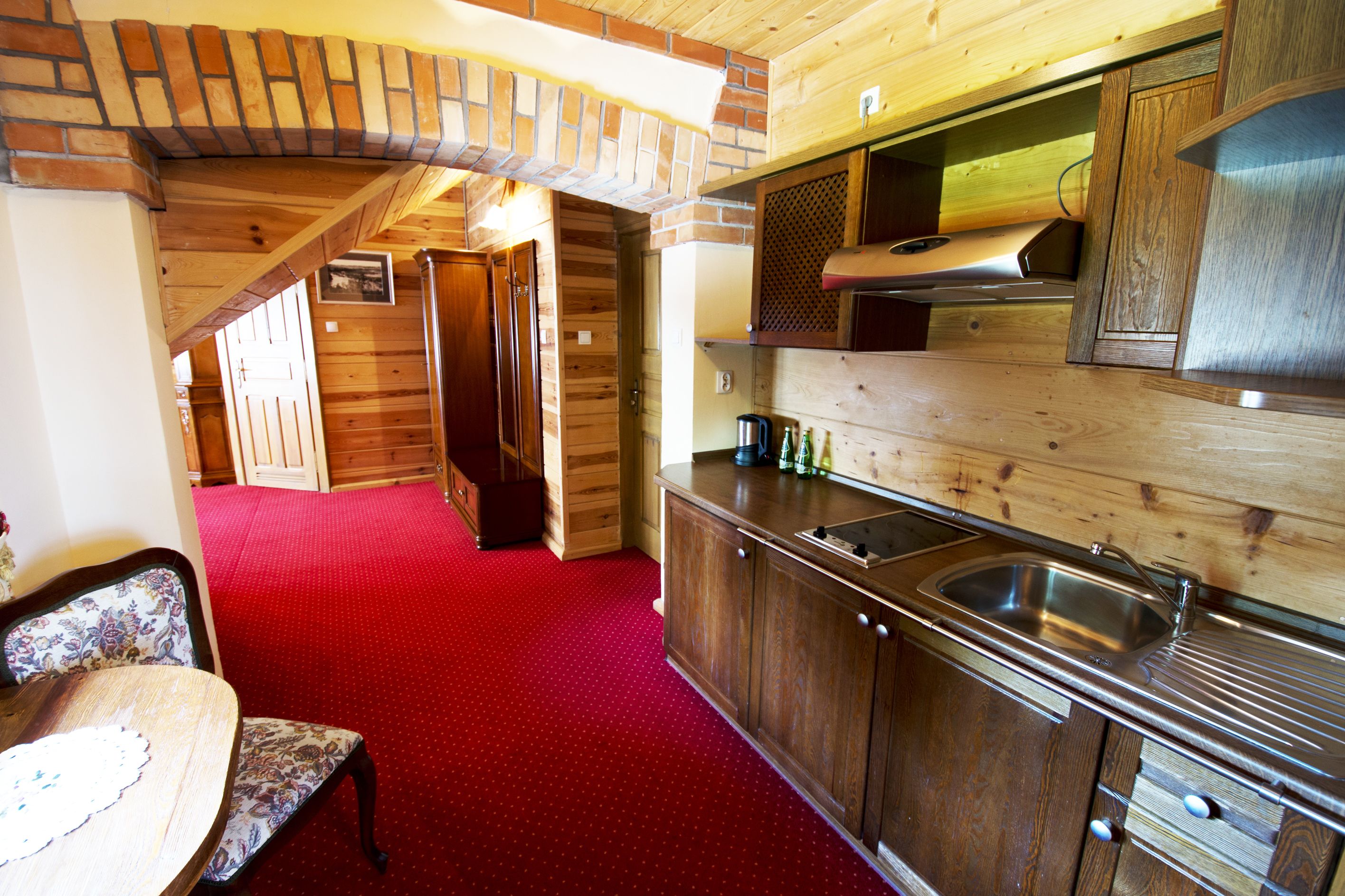 A wooden interior with a red carpet, a dining table with chairs, a kitchen area with a sink, and a wooden ceiling.