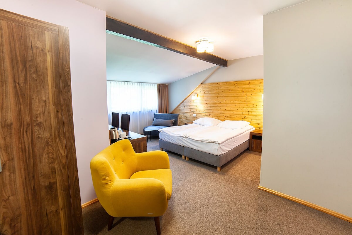 Une chambre à coucher avec un lit, une chaise jaune, un bureau avec une bouilloire et une fenêtre avec des rideaux.