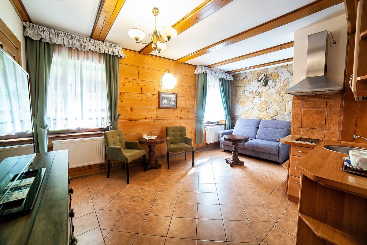 Une pièce chaleureuse avec des murs en bois, un mur d’accent en pierre, un canapé, deux fauteuils, une table à manger et un coin cuisine.