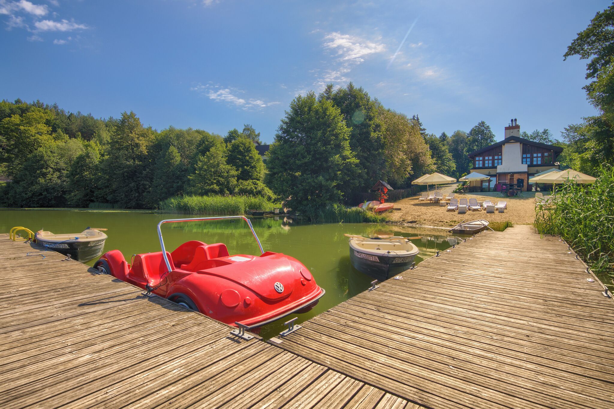 A wooden dock with a red boat, a house with umbrellas, and a lake surrounded by trees.