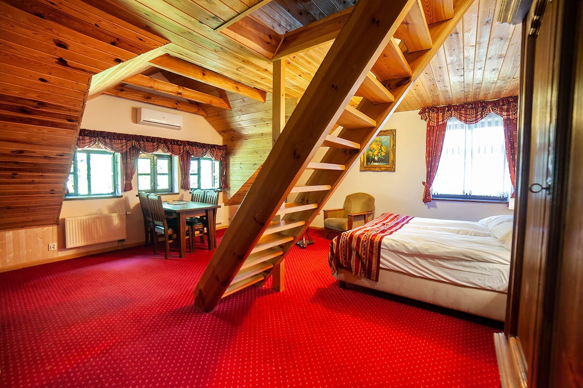 Une pièce avec un plafond en pente en bois, une moquette rouge, un lit, une table avec des chaises et une fenêtre avec des rideaux.