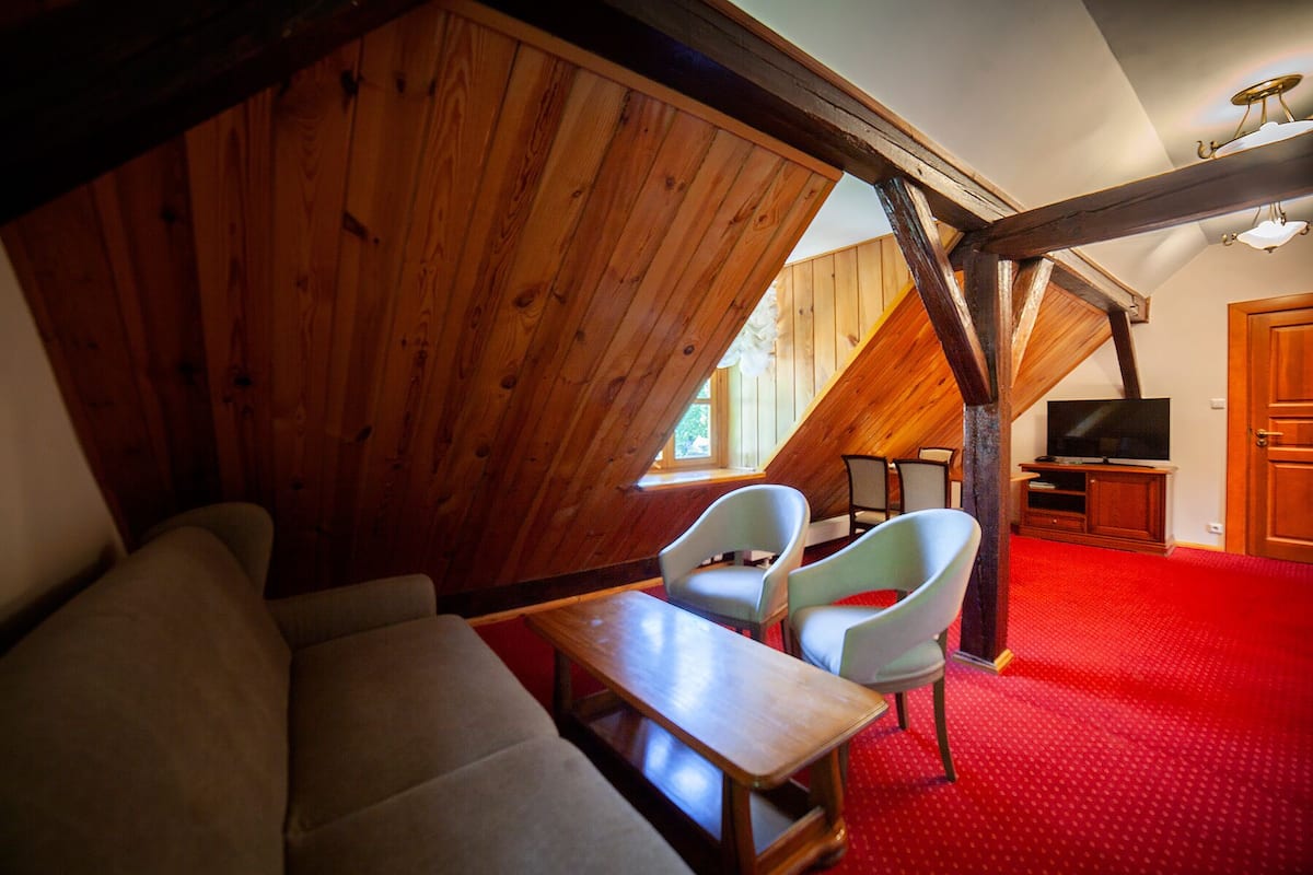 Une pièce mansardée chaleureuse avec des murs en bois, une moquette rouge, un canapé, deux fauteuils, un téléviseur et un meuble en bois.