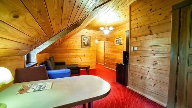 Interior acogedor de una cabaña con techo inclinado, paredes de madera, un comedor con mesa y sillas, y una pequeña zona de estar con cojines.