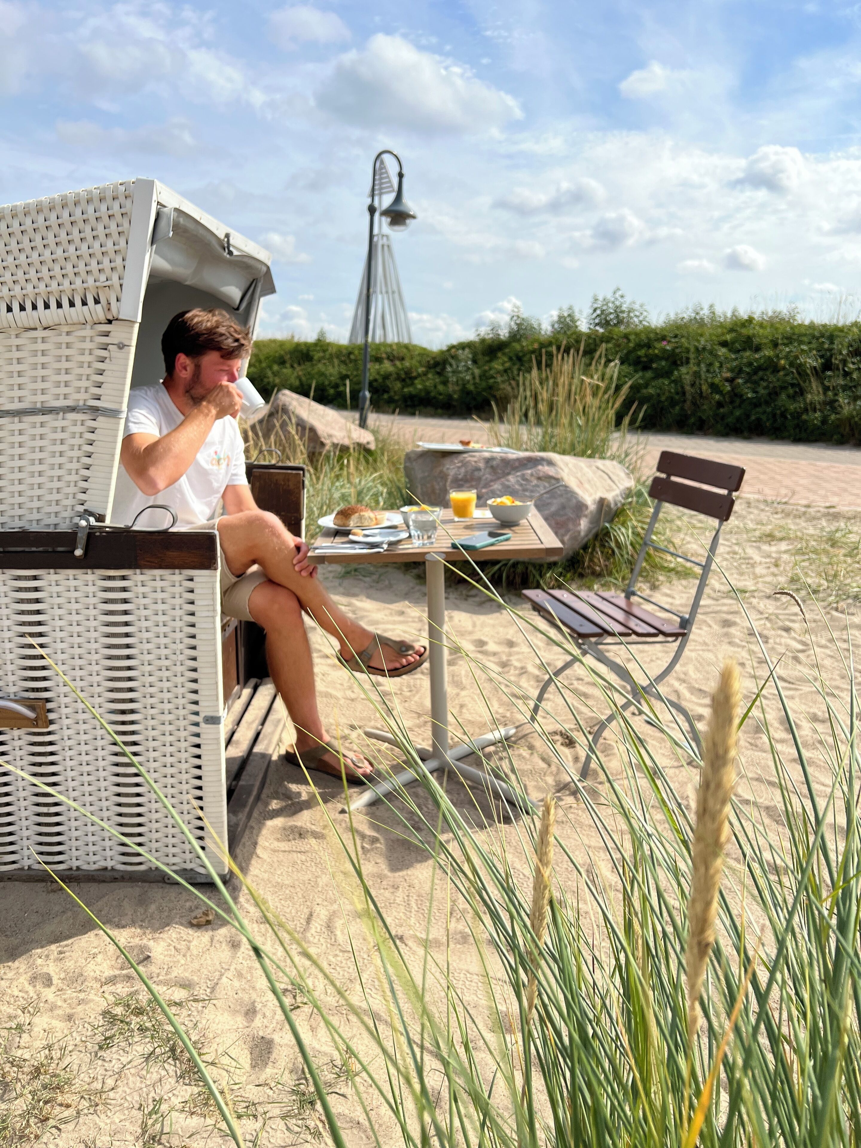 A beachside setting with a wicker lounge chair, a table with breakfast items, and a metal lamp post.